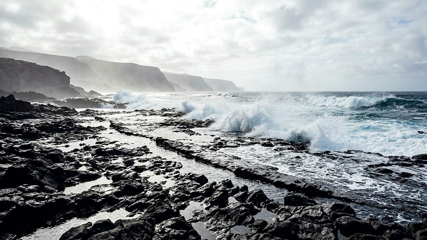 A rocky shoreline of dark basalt with waves breaking white across a low shelf under bright sky